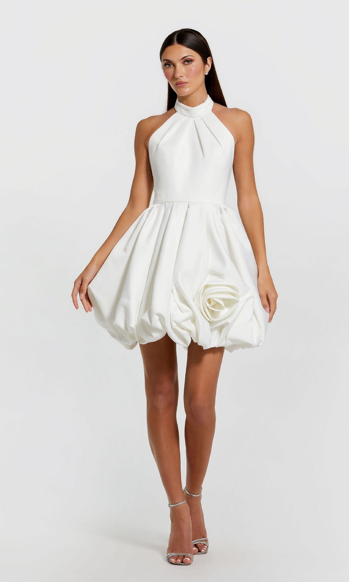 A woman wears the Mac Duggal 20707 Short Bubble-Hem Party Dress, a sleeveless satin halter style with a voluminous skirt and fabric rose detail, paired with silver high-heeled sandals.